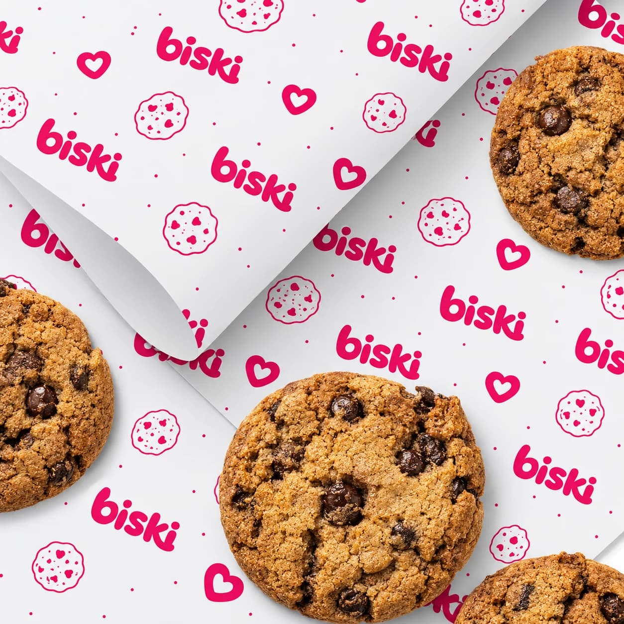 Cookies com gotas de chocolate sobre o papel de embrulho da biski, estampado com a marca, corações e cookies.
