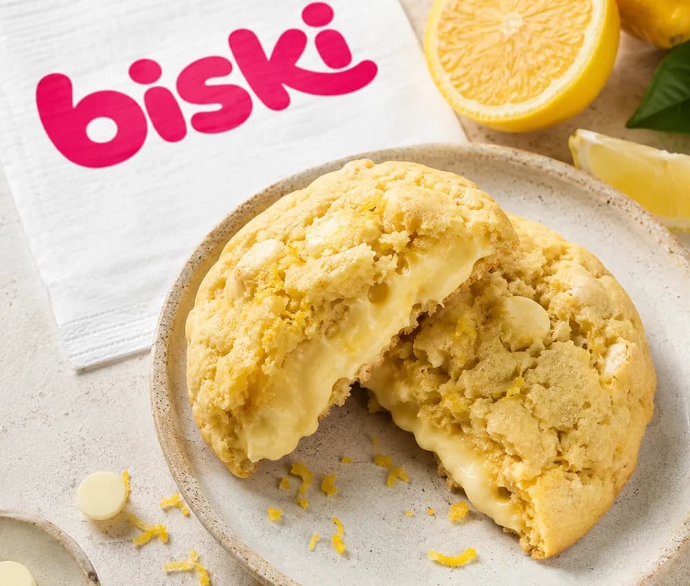 Cookie amarelo de limão siciliano aberto ao meio, com recheio cremoso claro, ao lado de limões cortados e raspas.
